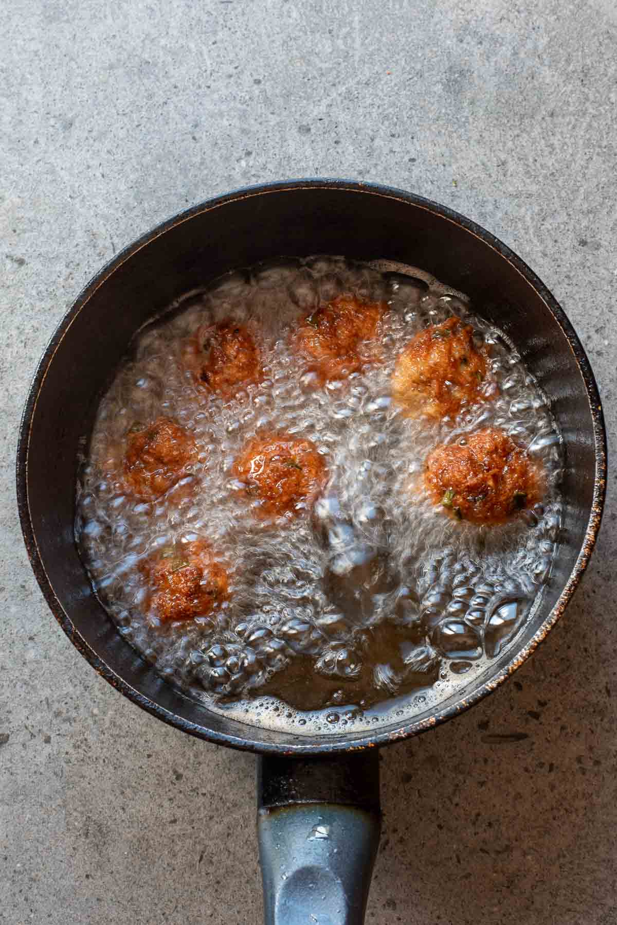 Six round fritters are being deep-fried in hot oil inside a black saucepan on a gray surface.