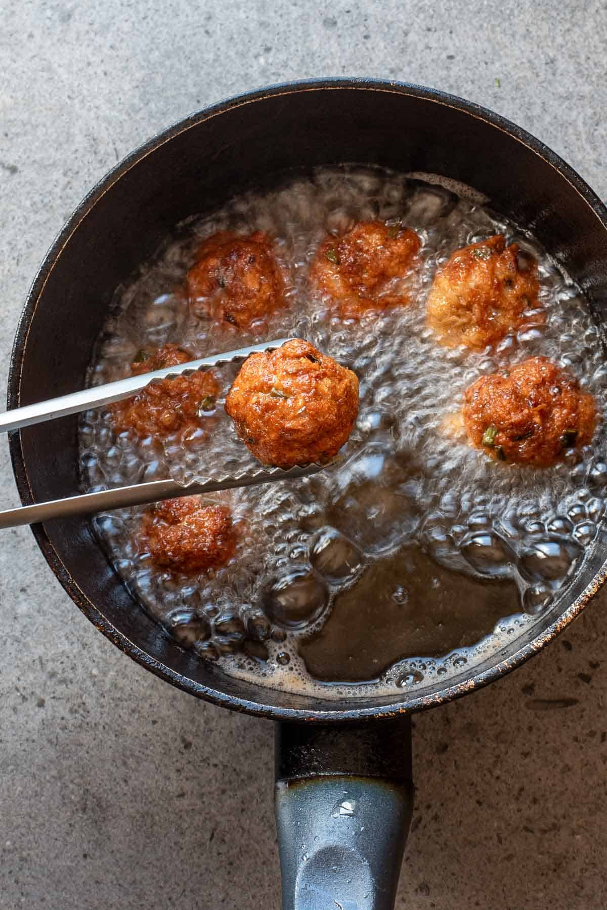 A close-up of several meatballs being deep-fried in a pot of oil, with tongs lifting one meatball out of the oil.