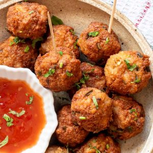 A plate of golden-brown fried meatballs garnished with chopped herbs, served with a side of red dipping sauce in a small bowl.