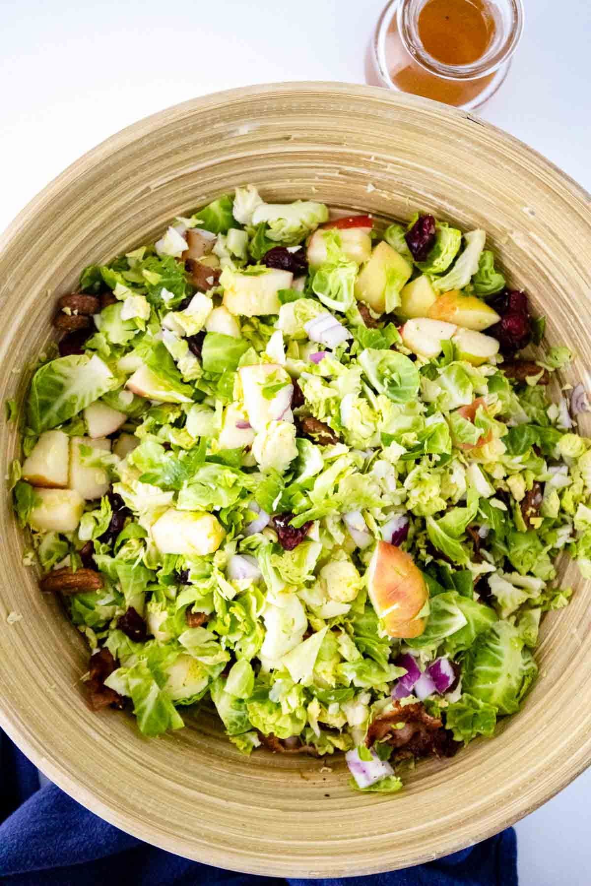 A wooden bowl filled with chopped salad featuring Brussels sprouts, apples, red onion, nuts, and dried cranberries sits beside a jar of dressing—perfect inspiration for fresh recipes.
