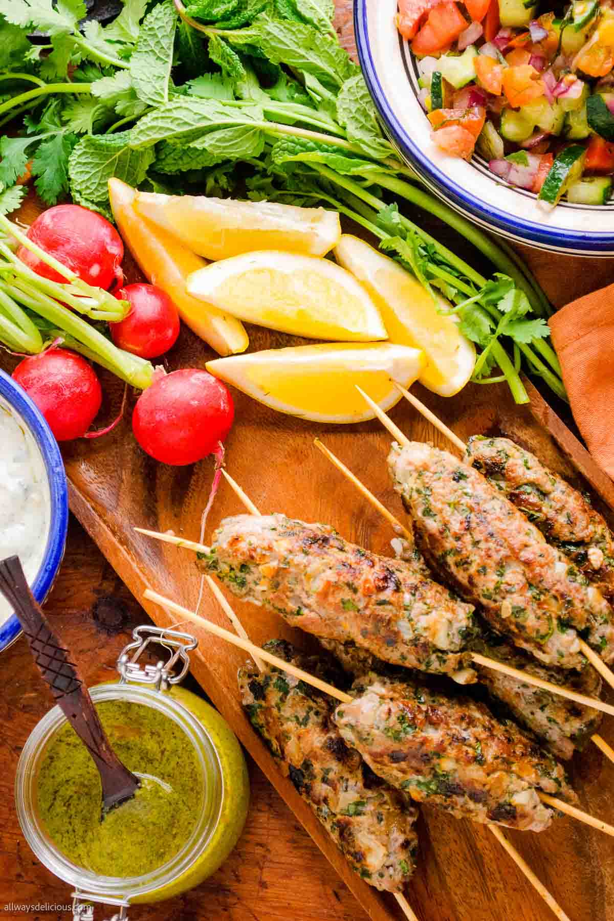 Grilled herb chicken skewers on a wooden board with lemon wedges, fresh radishes, greens, a bowl of chopped salad, and a jar of green sauce—perfect for adding to your collection of summer recipes.