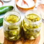 Two glass jars filled with pickled jalapenos sit on a wooden surface, surrounded by fresh jalapeños, a bowl of salt, and jar lids in the background.