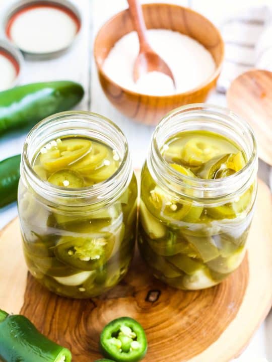 Two glass jars filled with pickled jalapenos sit on a wooden surface, surrounded by fresh jalapeños, a bowl of salt, and jar lids in the background.