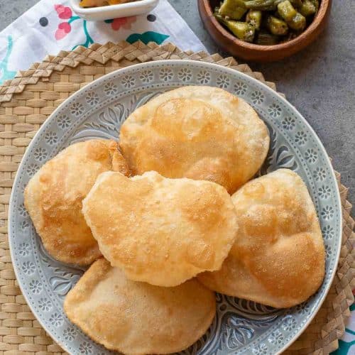 A plate of five puffed puris, served with a small bowl of mango pickle and a bowl of cooked okra, on a woven mat.