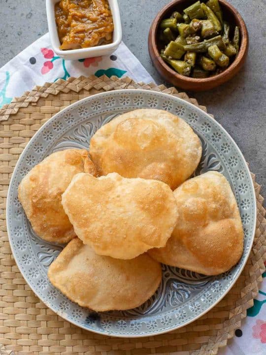 A plate of five puffed puris, served with a small bowl of mango pickle and a bowl of cooked okra, on a woven mat.