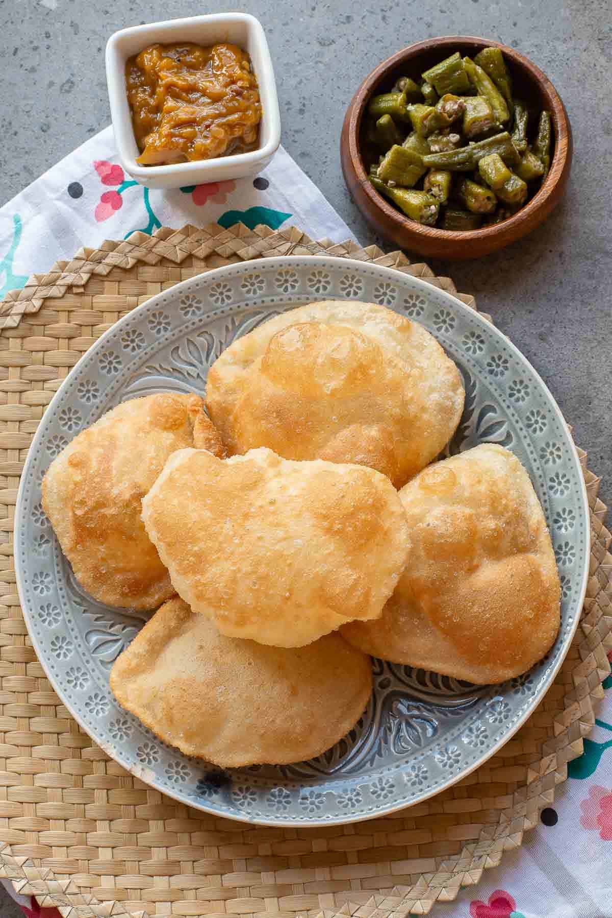 A plate of five puffed puris, served with a small bowl of mango pickle and a bowl of cooked okra, on a woven mat.