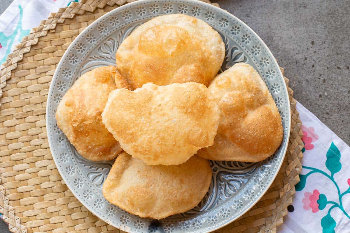 A plate with five pieces of golden, puffy fried bread from a classic poori recipe sits on a woven placemat with a floral-patterned cloth nearby.