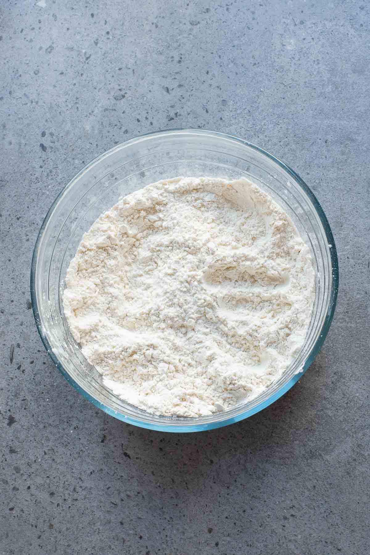 A glass bowl filled with flour and other dry ingredients sits on a gray countertop.
