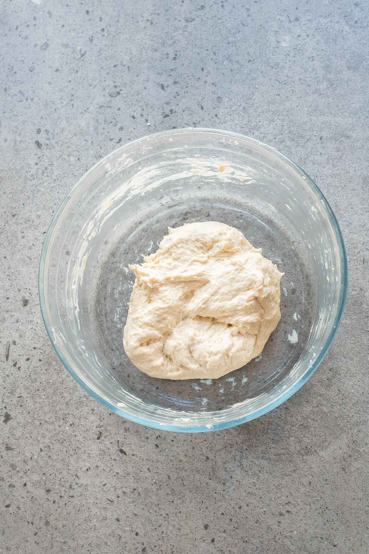 A clear glass bowl containing a portion of raw bread dough rests on a gray speckled countertop.