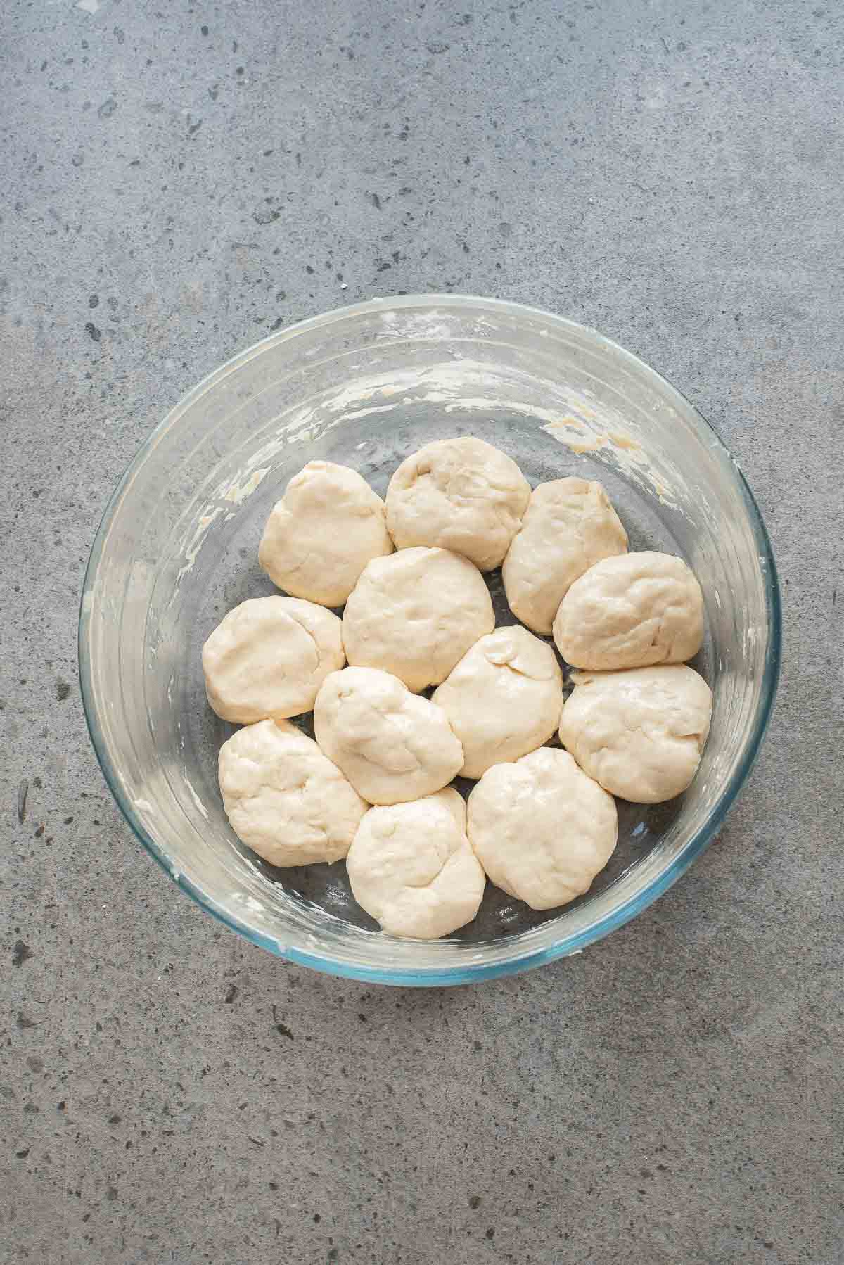 A glass bowl containing twelve round pieces of raw dough on a gray countertop.