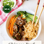 A bowl of Chinese beef and noodle soup with bok choy, carrots, and scallions, served with chopsticks. A small bowl of scallions and a jar of chili oil are nearby.