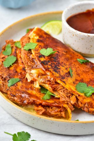 Two folded birria tacos filled with shredded meat and cheese, garnished with cilantro, served on a plate with a bowl of dipping sauce and a lime wedge.