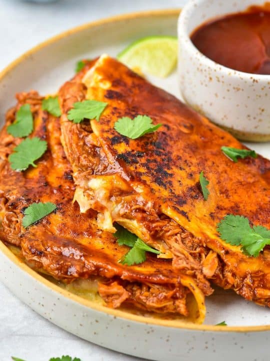 Two folded birria tacos filled with shredded meat and cheese, garnished with cilantro, served on a plate with a bowl of dipping sauce and a lime wedge.