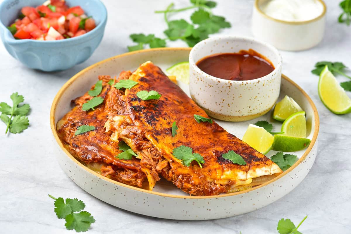 Two cheesy quesadillas with shredded meat on a plate, garnished with cilantro, served with lime wedges, red sauce, sour cream, and a bowl of pico de gallo.