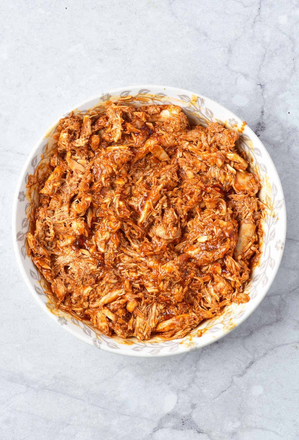 A white bowl filled with shredded meat mixed with a reddish-brown sauce, placed on a marble surface.