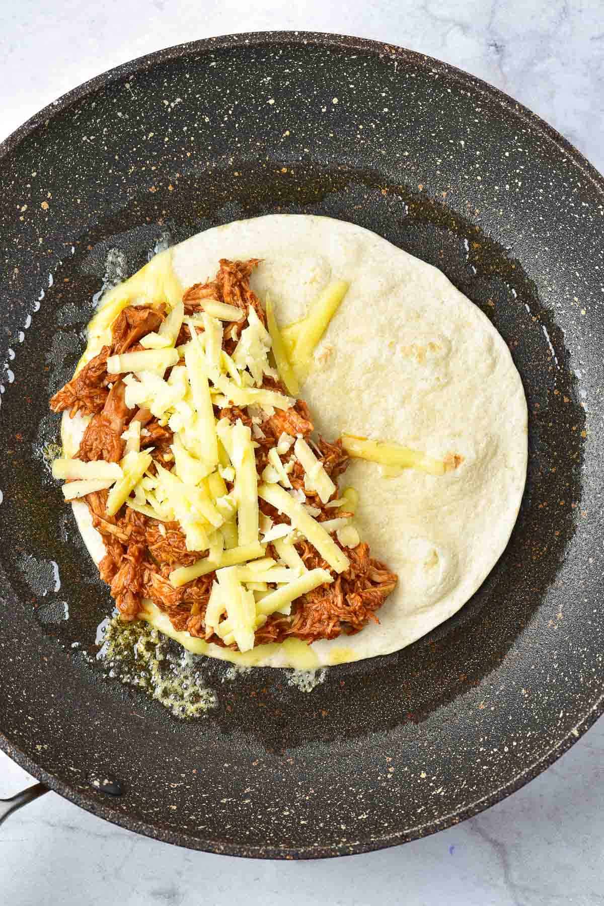 A flour tortilla in a skillet is topped with shredded meat and grated cheese, ready to be folded and cooked.