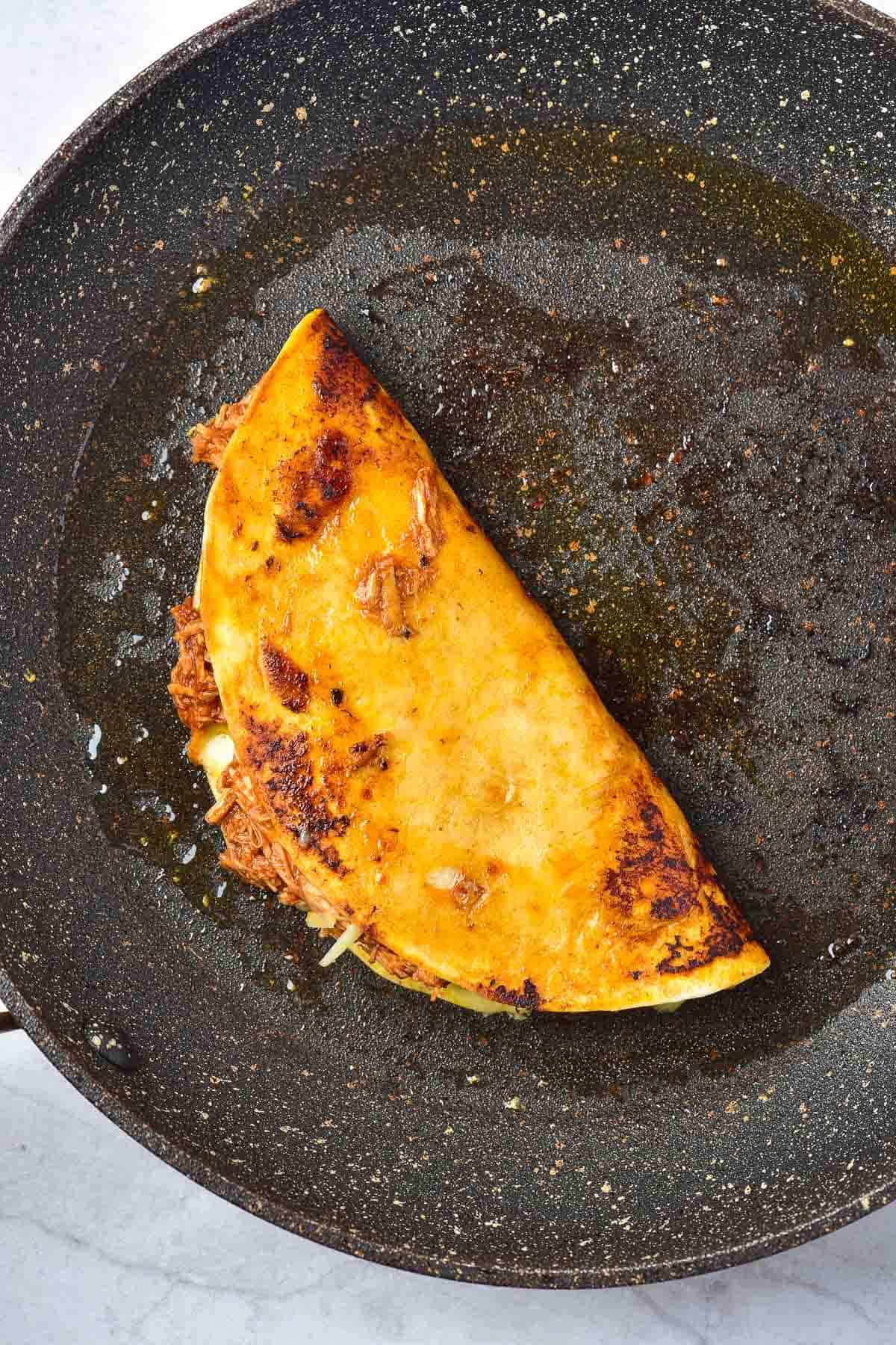 A folded, browned taco filled with shredded meat and cheese is cooking in a lightly oiled skillet.