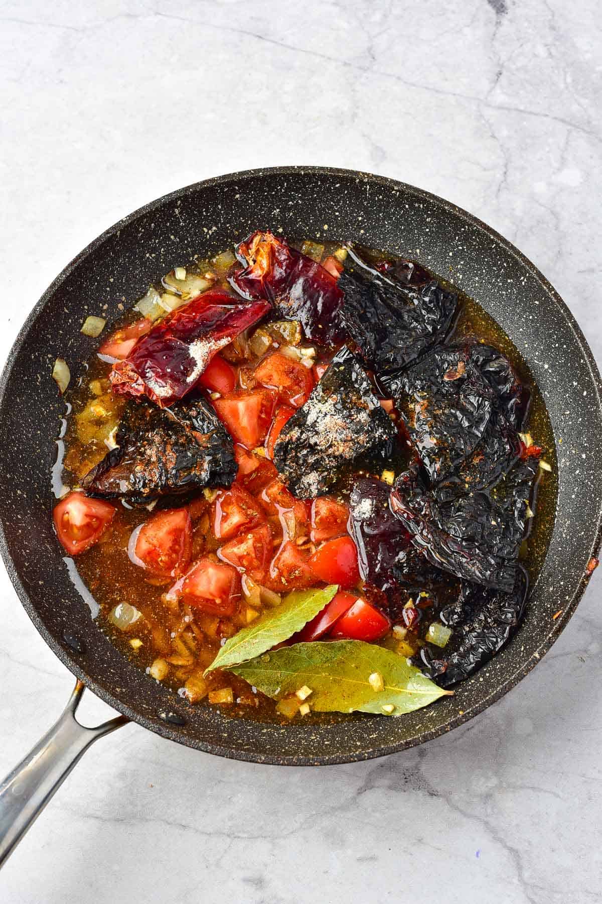 A frying pan contains chopped tomatoes, dried chili peppers, bay leaves, onions, and liquid, sitting on a light-colored countertop.