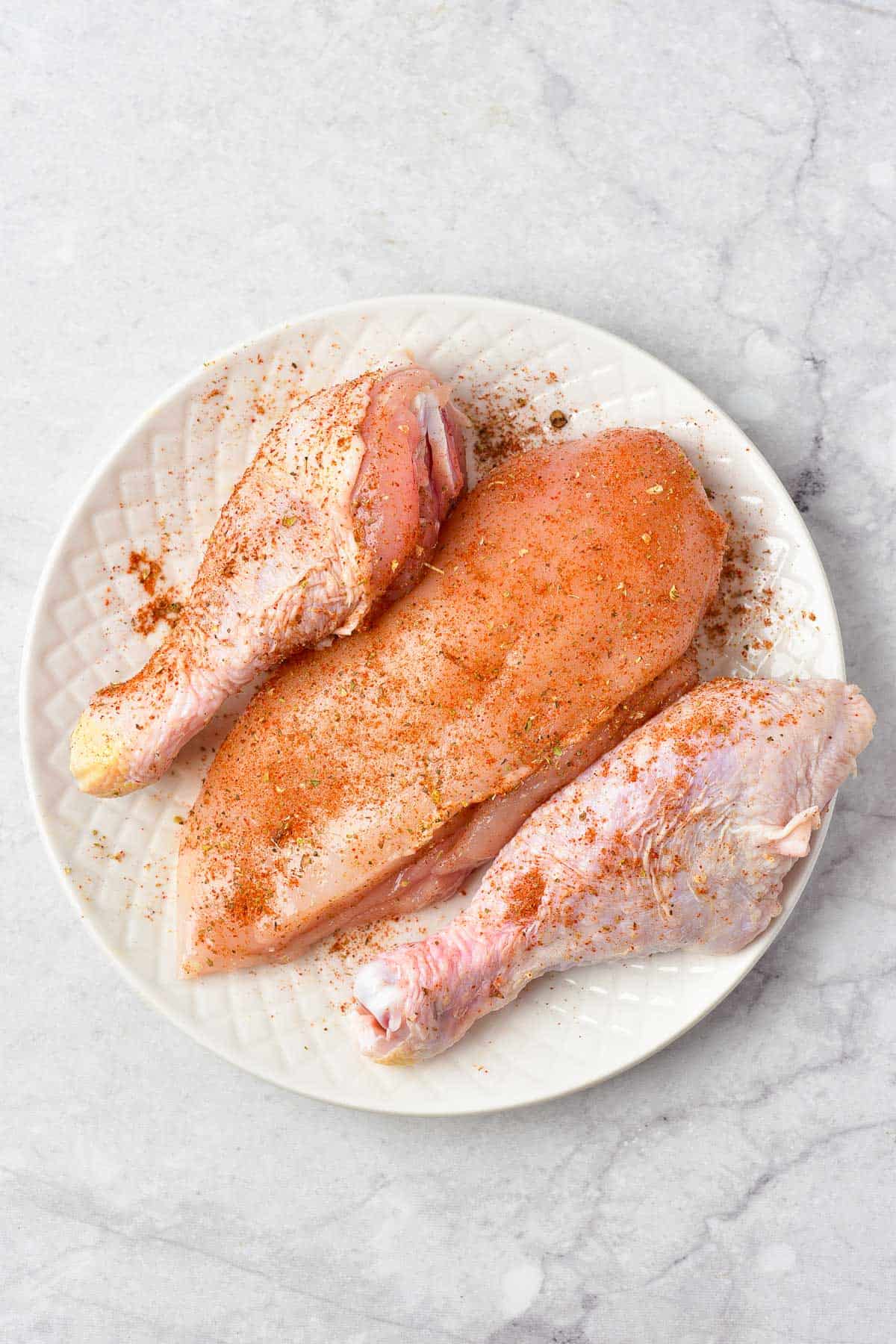 A plate with two raw chicken drumsticks and one raw chicken breast, all sprinkled with seasoning, on a light marble surface.