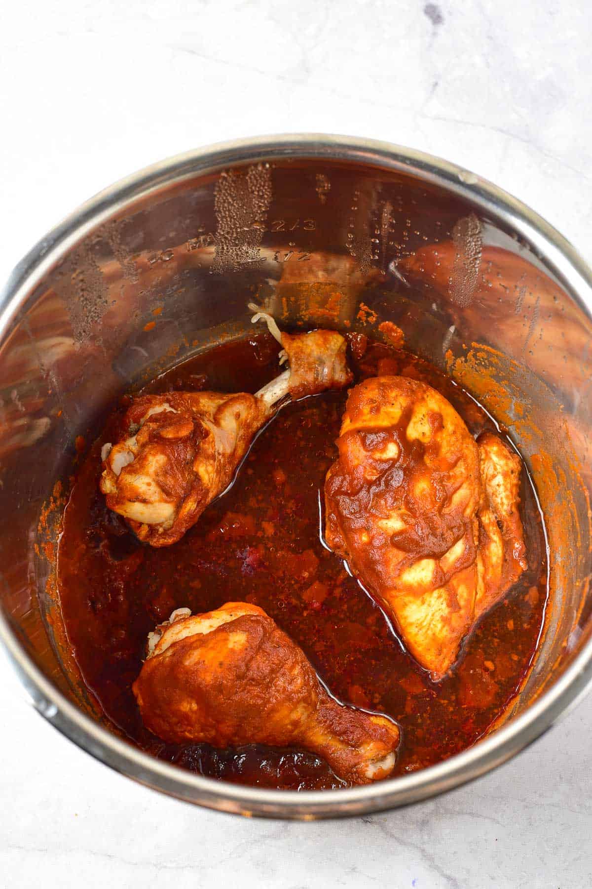 Three pieces of seasoned chicken in a reddish sauce inside a stainless steel pot on a marble surface.