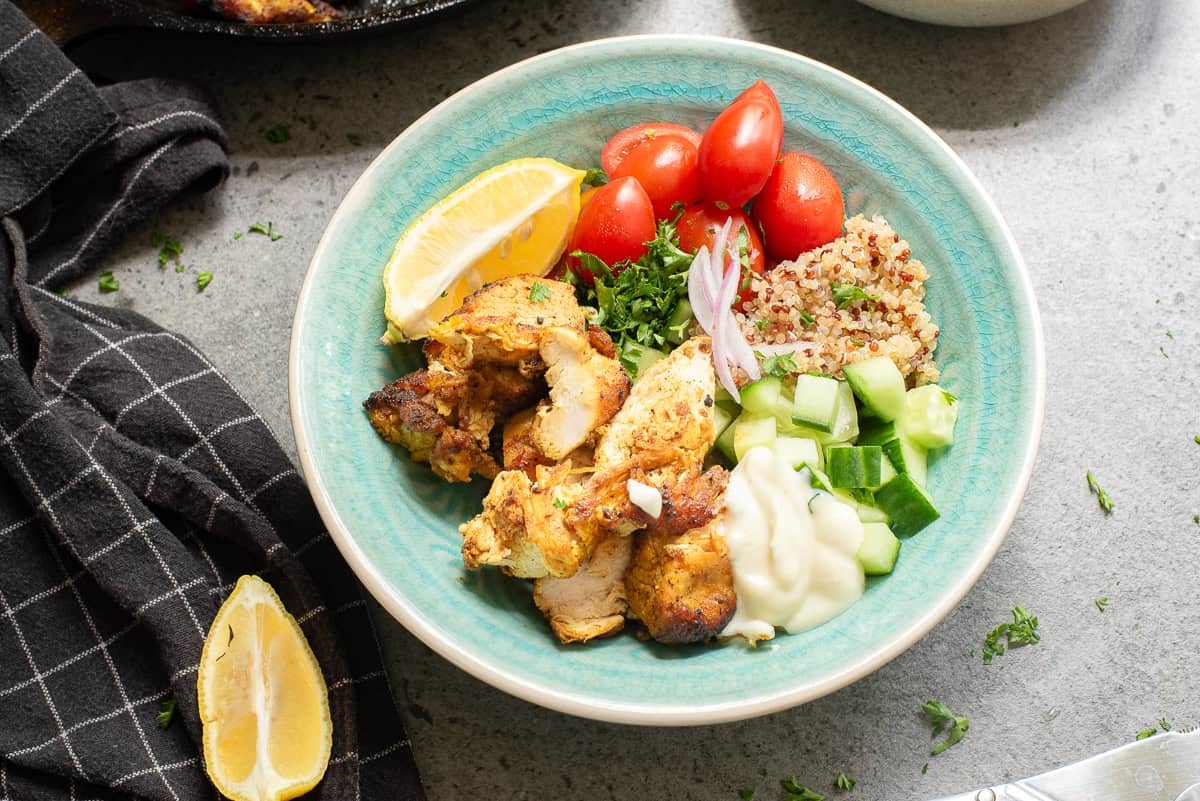 A bowl with grilled chicken, quinoa, cucumber, grape tomatoes, fresh parsley, sliced onion, a lemon wedge, and a dollop of white sauce on a gray surface with a black towel nearby.