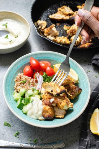 A hand holds a fork over a bowl of grilled chicken, diced cucumber, cherry tomatoes, quinoa, red onion, and yogurt sauce, with a skillet and sauce bowl nearby.