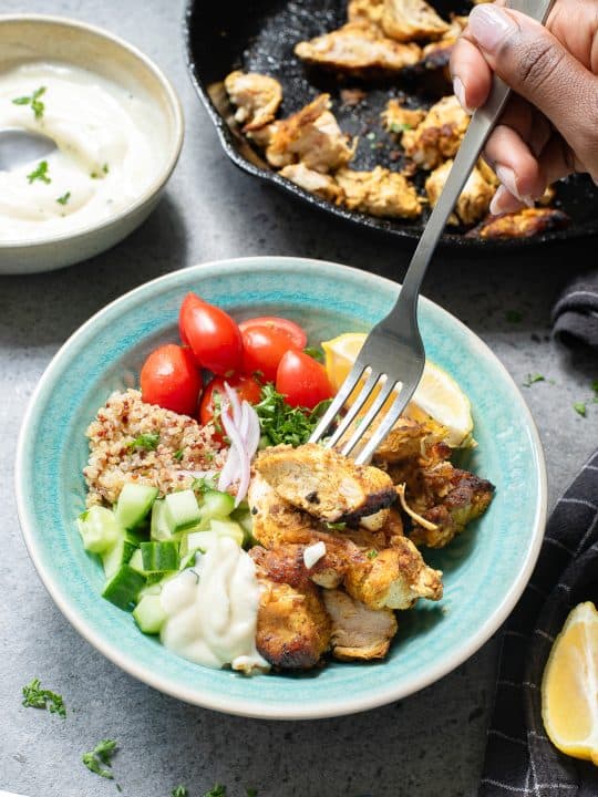 A hand holds a fork over a bowl of grilled chicken, diced cucumber, cherry tomatoes, quinoa, red onion, and yogurt sauce, with a skillet and sauce bowl nearby.
