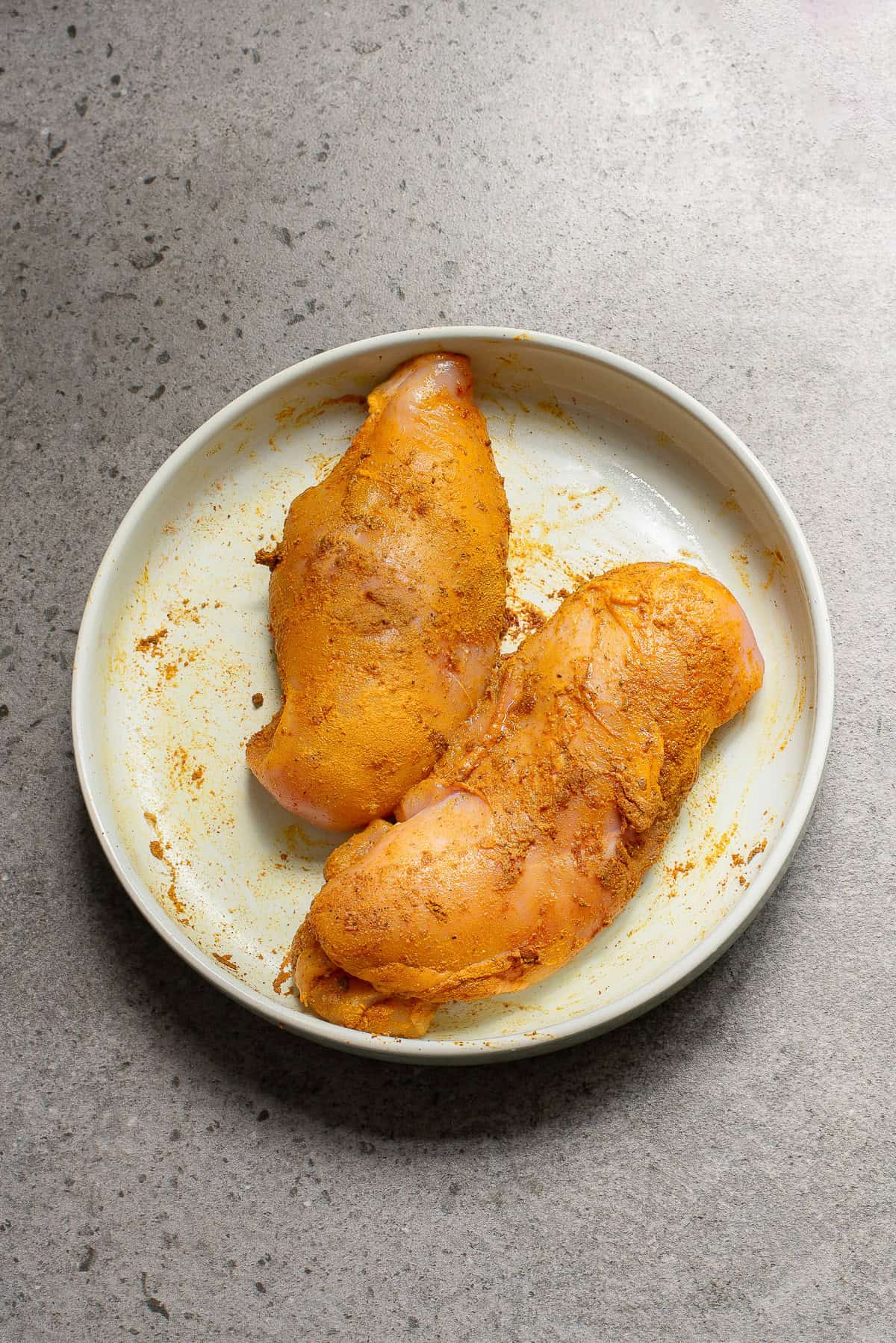 Two raw, seasoned chicken breasts on a white plate placed on a gray, speckled countertop.
