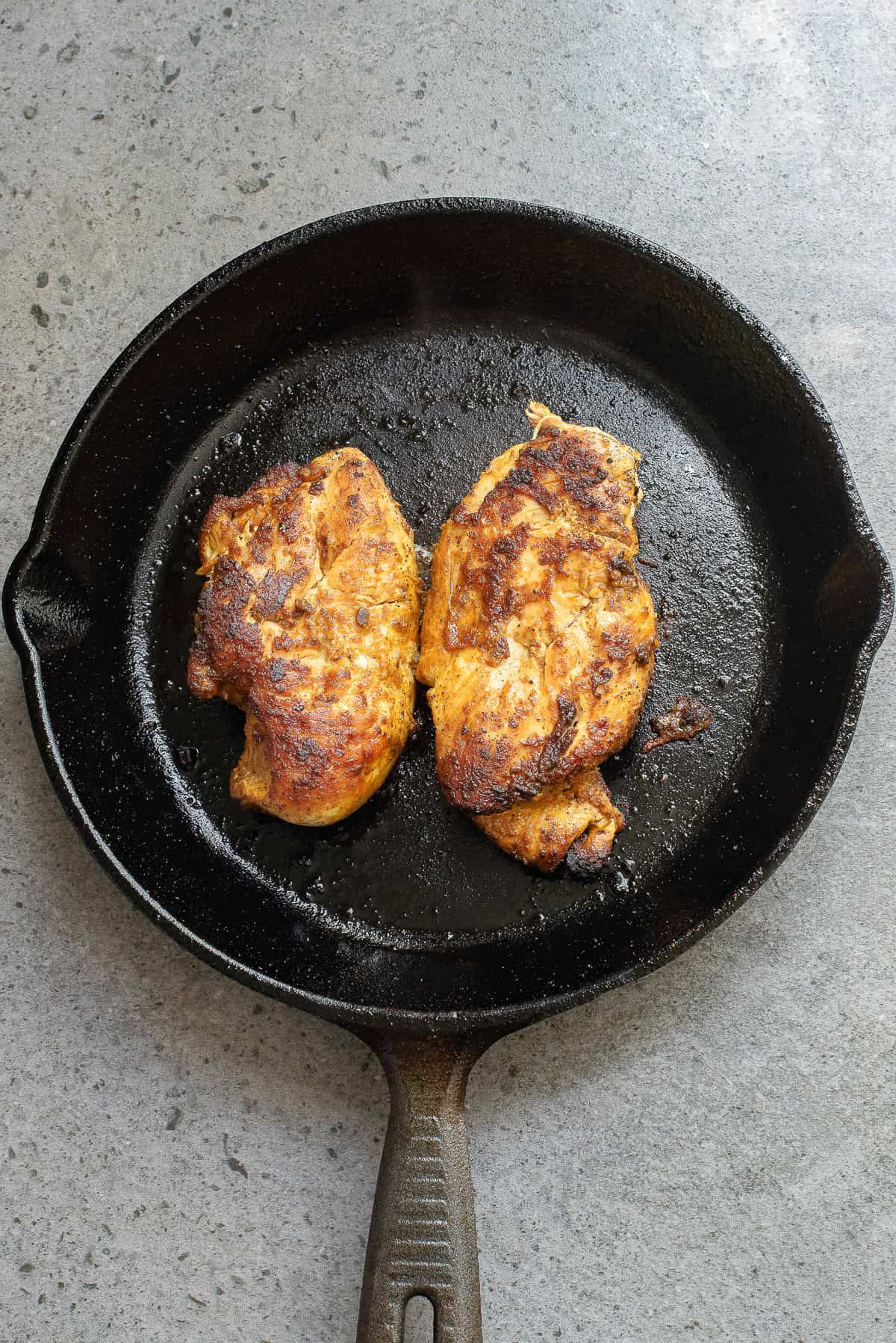 Two cooked, seasoned chicken breasts in a black cast iron skillet on a gray countertop.