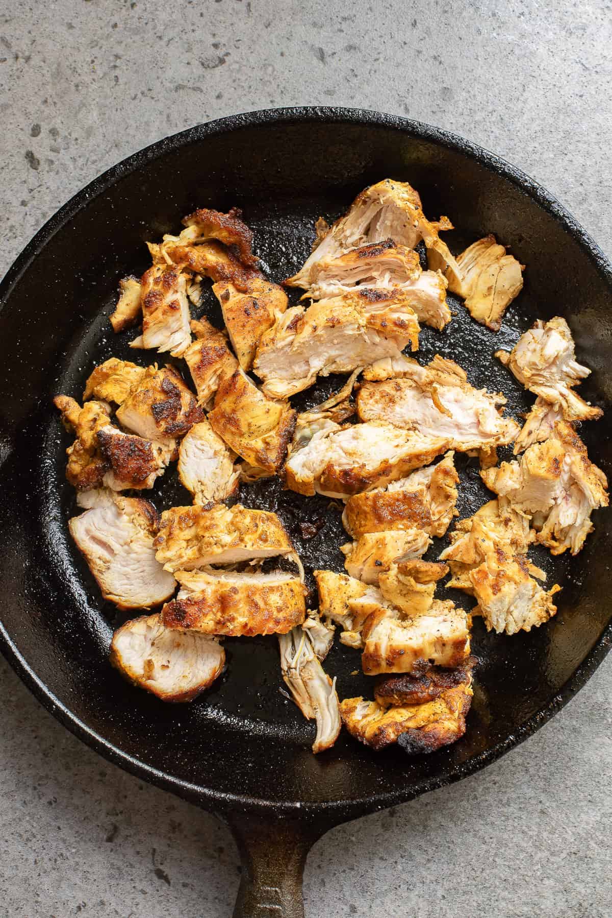 Cooked, sliced seasoned chicken pieces in a black cast iron skillet on a gray textured surface.