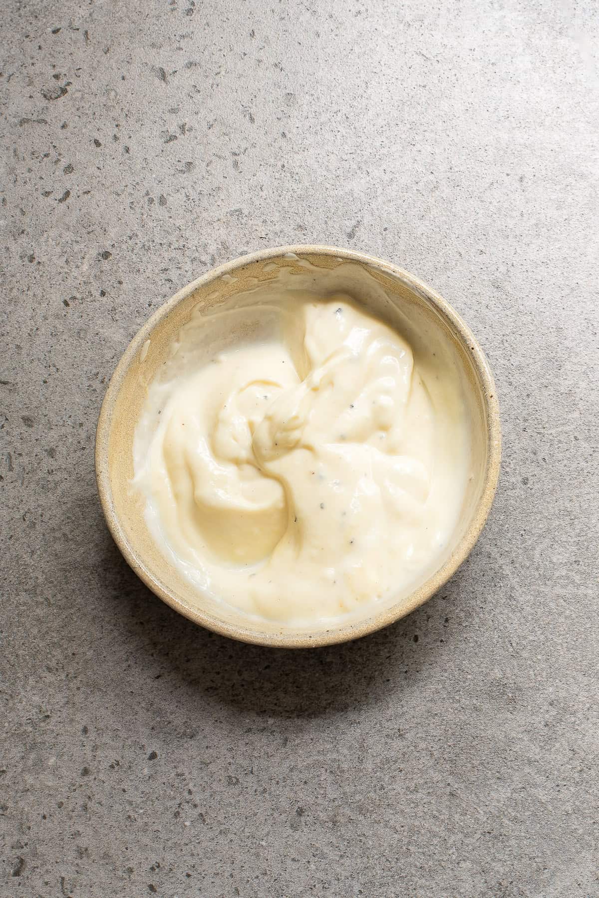 A beige bowl filled with creamy white sauce or dip sits on a gray textured surface.