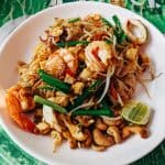 A plate of pad thai with shrimp, tofu, bean sprouts, green onions, cashews, and a lime wedge on a green elephant-patterned tablecloth.