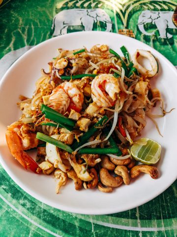 A plate of pad thai with shrimp, tofu, bean sprouts, green onions, cashews, and a lime wedge on a green elephant-patterned tablecloth.