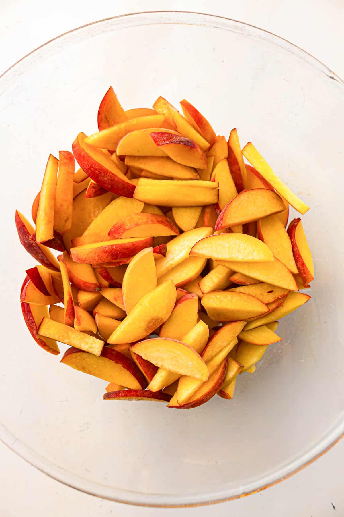 Sliced yellow peaches with red skins in a clear glass bowl on a white surface.
