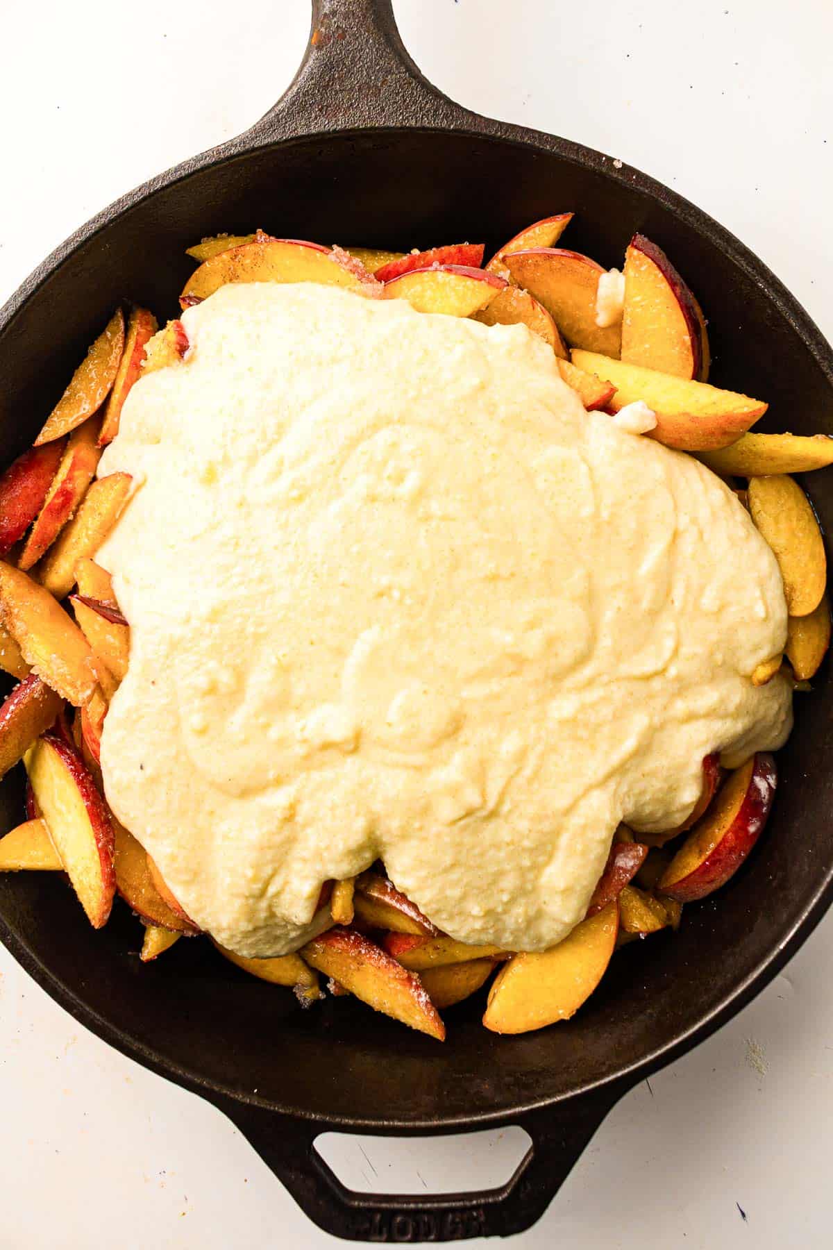 Sliced peaches in a cast iron skillet are topped with an uncooked layer of batter, ready to be baked.