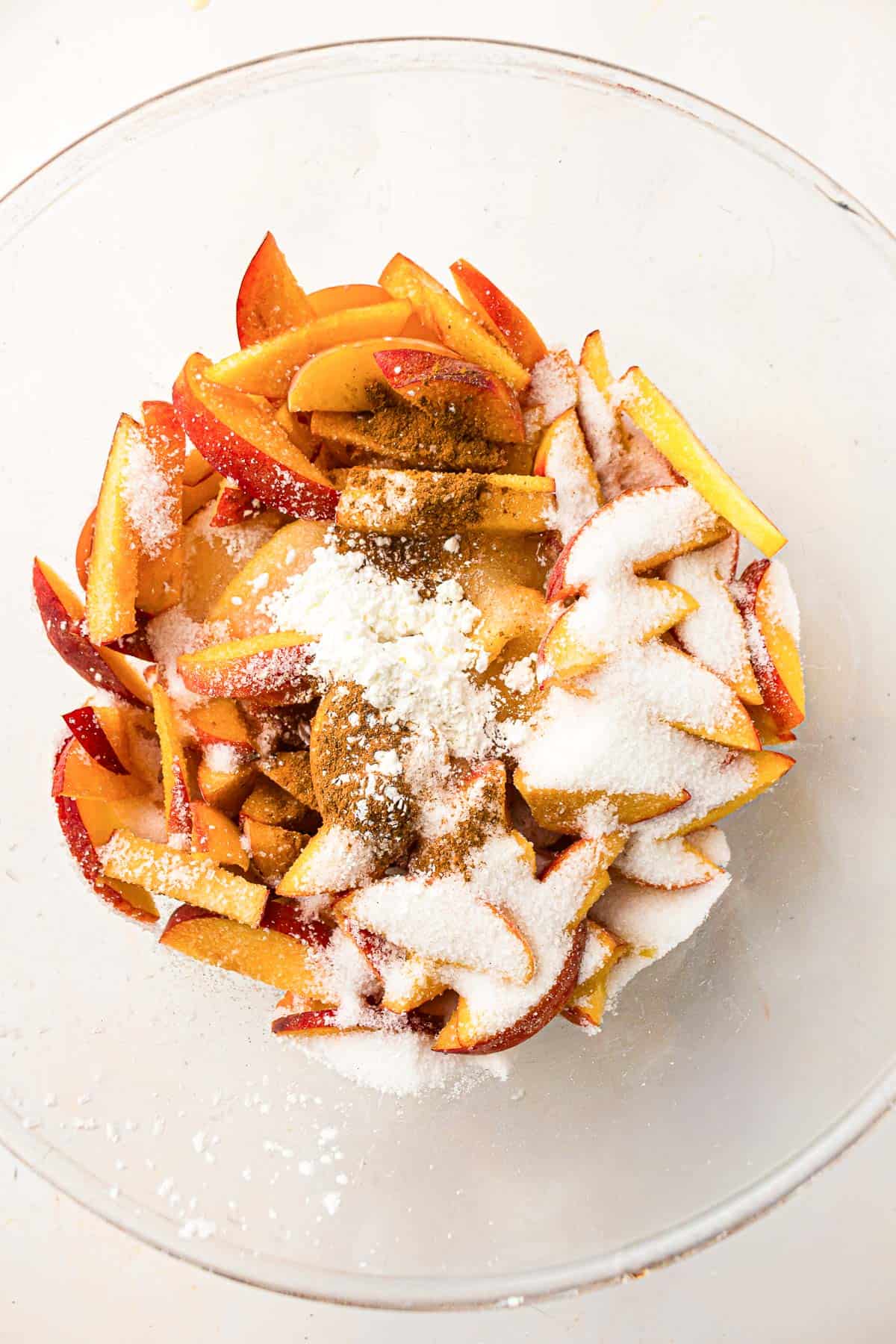 Sliced peaches in a glass bowl topped with sugar, spices, and cornstarch, ready to be mixed for baking.