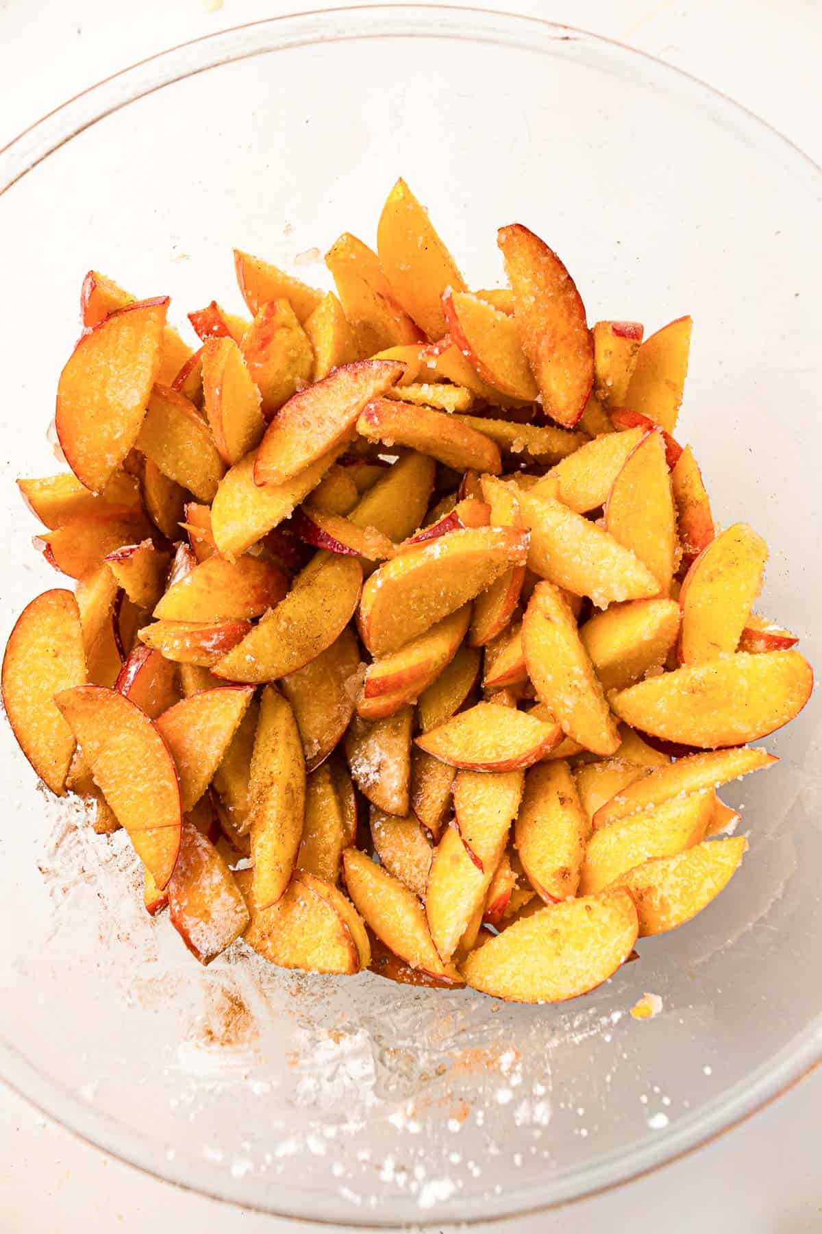 Sliced peaches mixed with sugar and spices in a clear glass bowl.