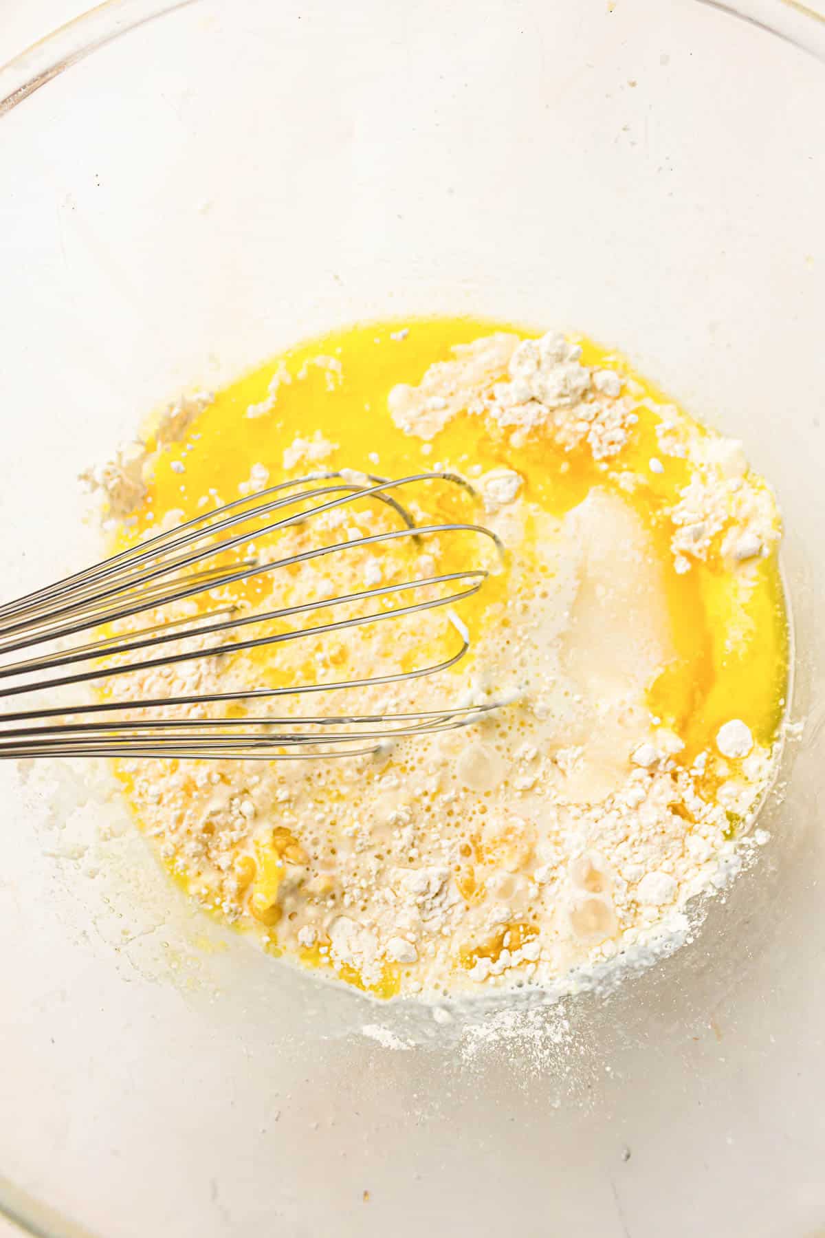 A metal whisk in a glass bowl with flour, sugar, eggs, and liquid ingredients being mixed together.