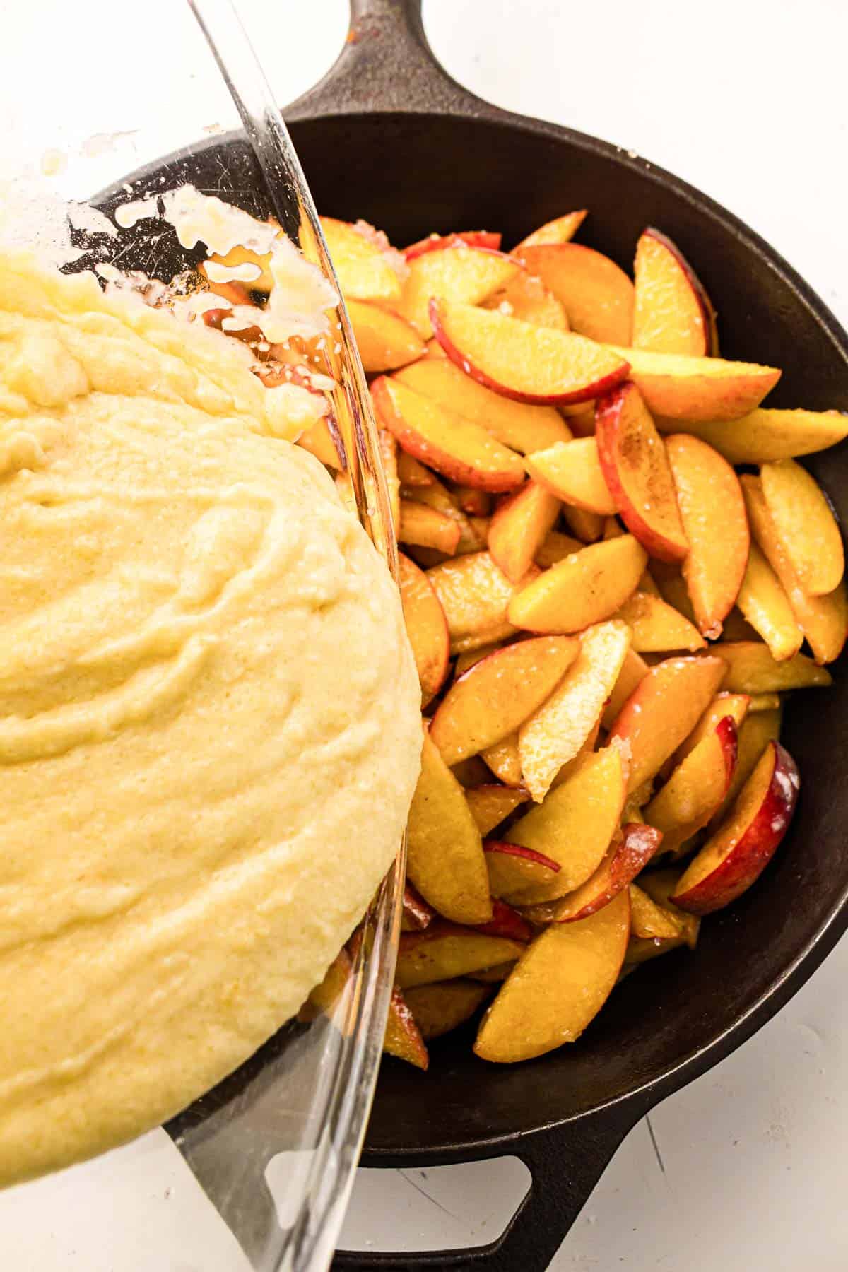 A bowl of thick batter is being poured over sliced peaches in a black cast iron skillet.