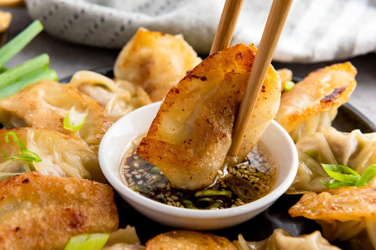 A pair of chopsticks holds a potsticker, dipping it into a small bowl of soy-based sauce with sesame seeds, surrounded by more dumplings on a plate.