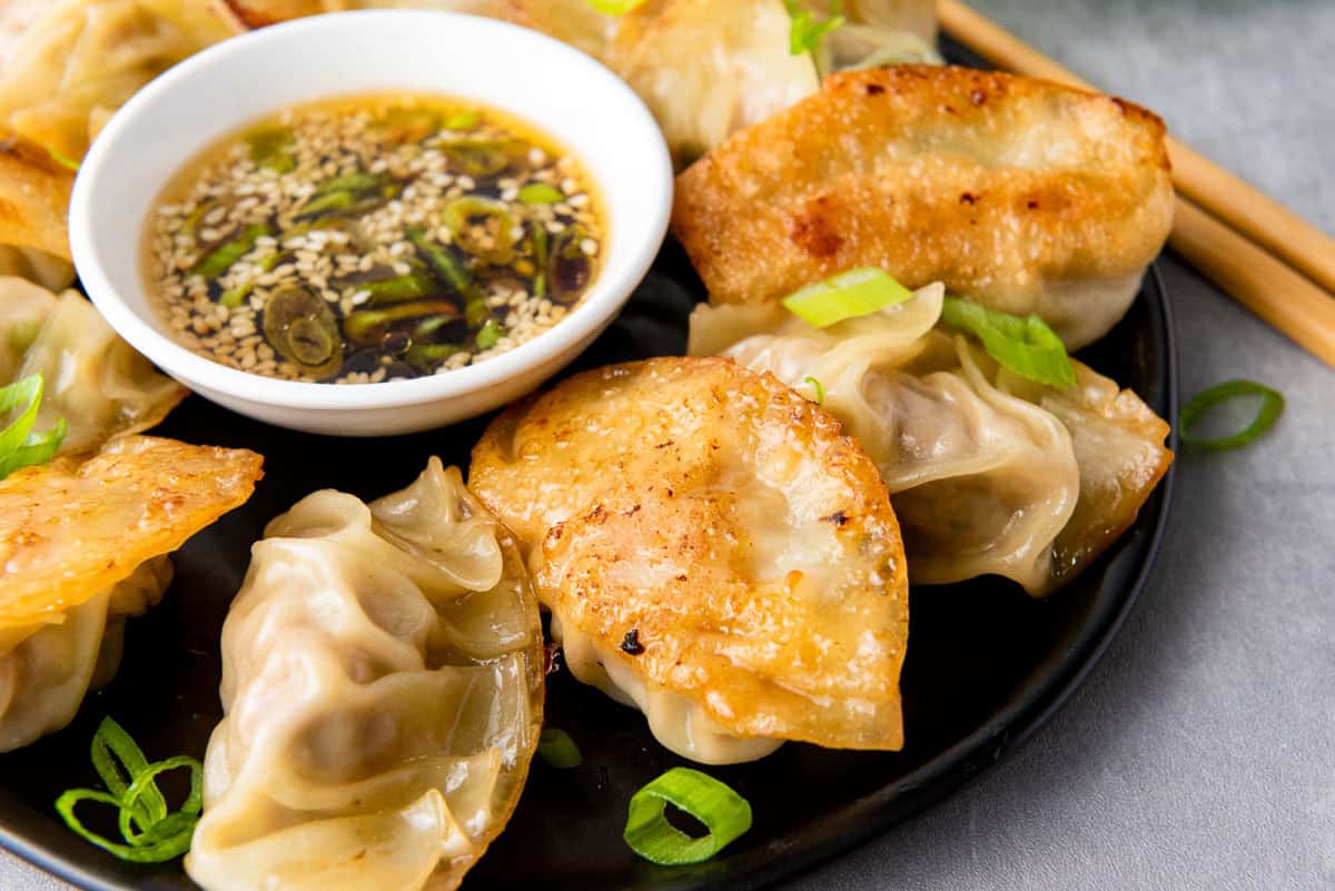 A plate of potstickers with chopped green onions, served with a bowl of soy dipping sauce topped with sliced green chilies and sesame seeds.
