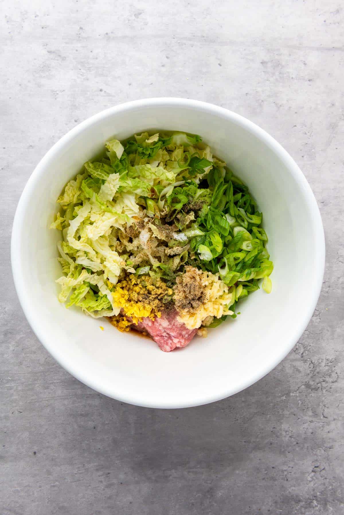 A white bowl contains chopped lettuce, green onions, ground meat, minced garlic, and various seasonings on a gray surface.