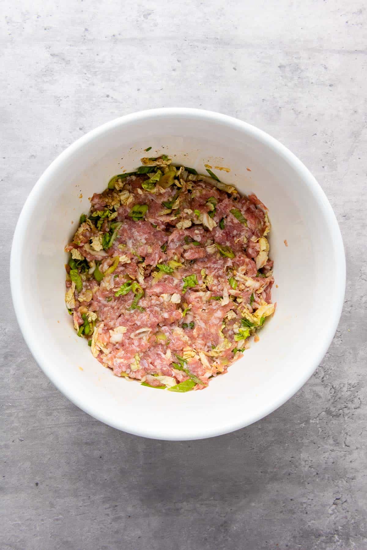 A white bowl containing a mixture of raw ground meat and chopped green vegetables on a light gray surface.