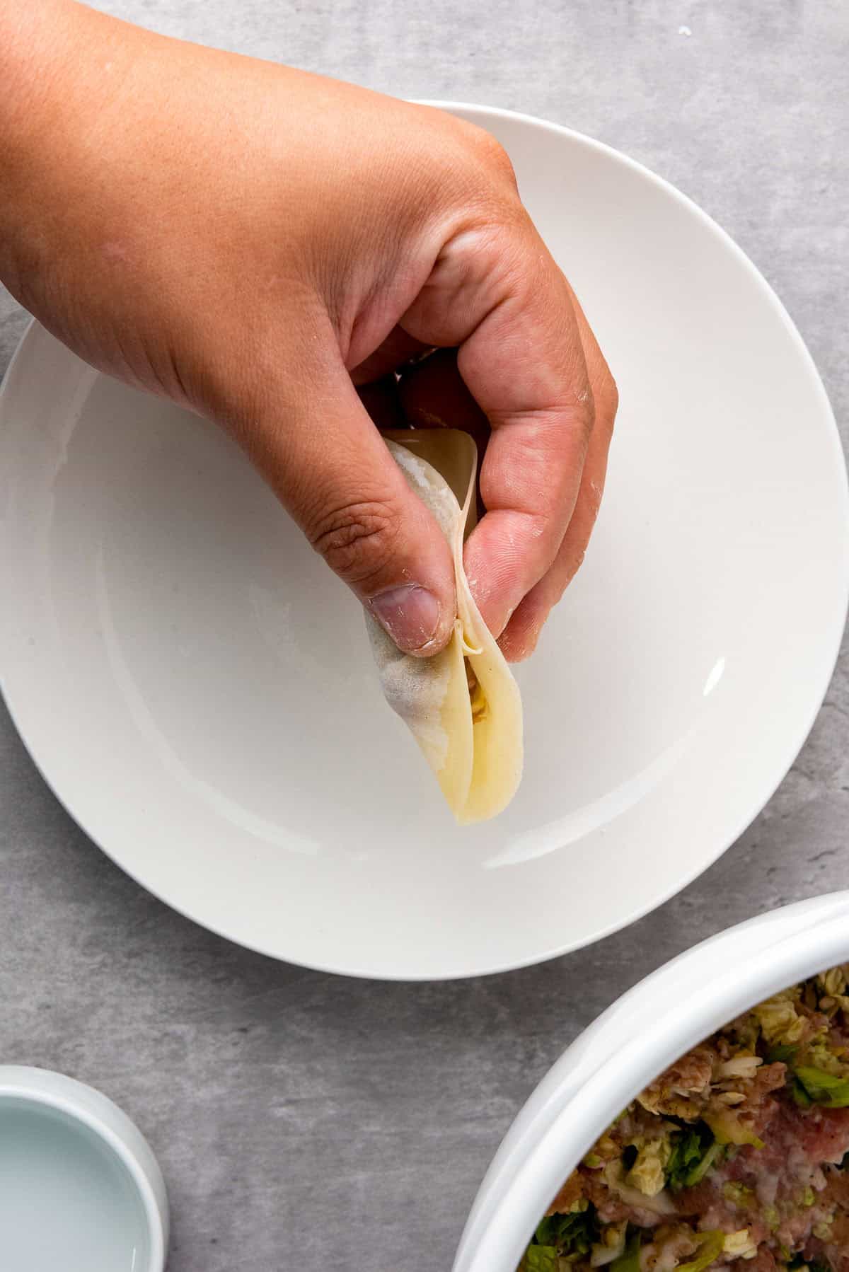 A hand is folding a dumpling wrapper with filling inside over a white plate, with a bowl of filling nearby on a gray surface.