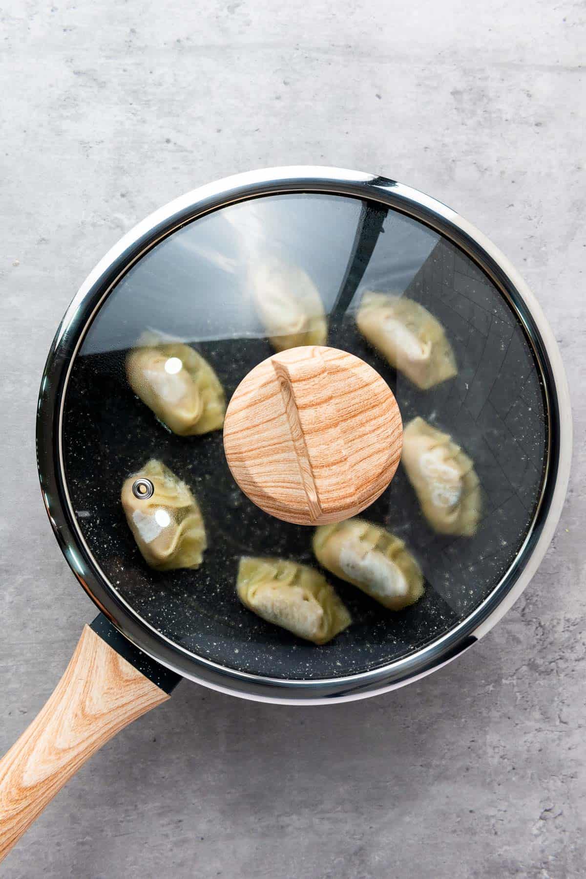 A frying pan with a glass lid containing eight dumplings is placed on a gray countertop.