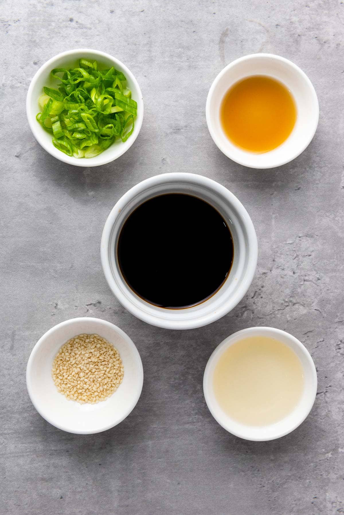 Five small white bowls containing chopped green onions, soy sauce, sesame seeds, rice vinegar, and sesame oil, arranged on a gray surface.
