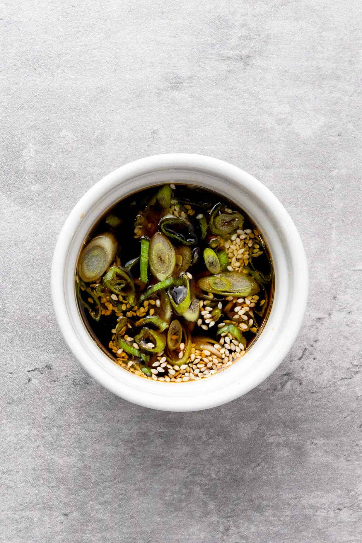 A small white bowl filled with soy sauce, sliced green onions, and sesame seeds on a light gray surface.