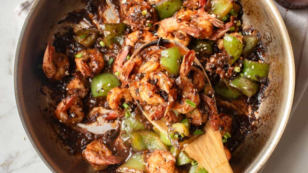 Shrimp and green bell peppers cooked in a dark sauce are being stirred in a skillet with a wooden spoon.