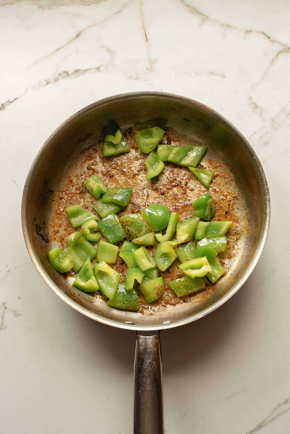 Chopped green bell pepper pieces cooking in a stainless steel pan on a light-colored marble surface.