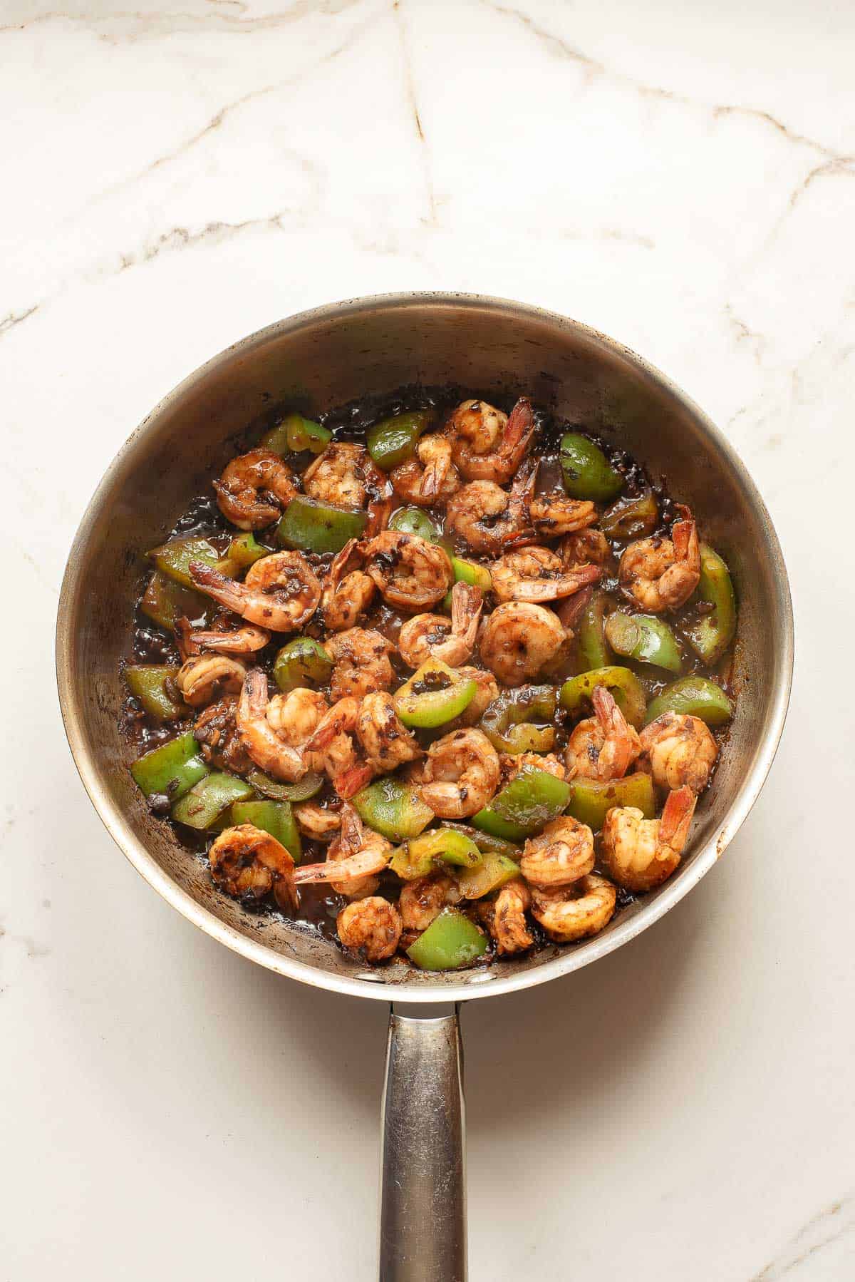 A stainless steel pan filled with cooked shrimp and chopped green bell peppers in a dark sauce on a white marble surface.